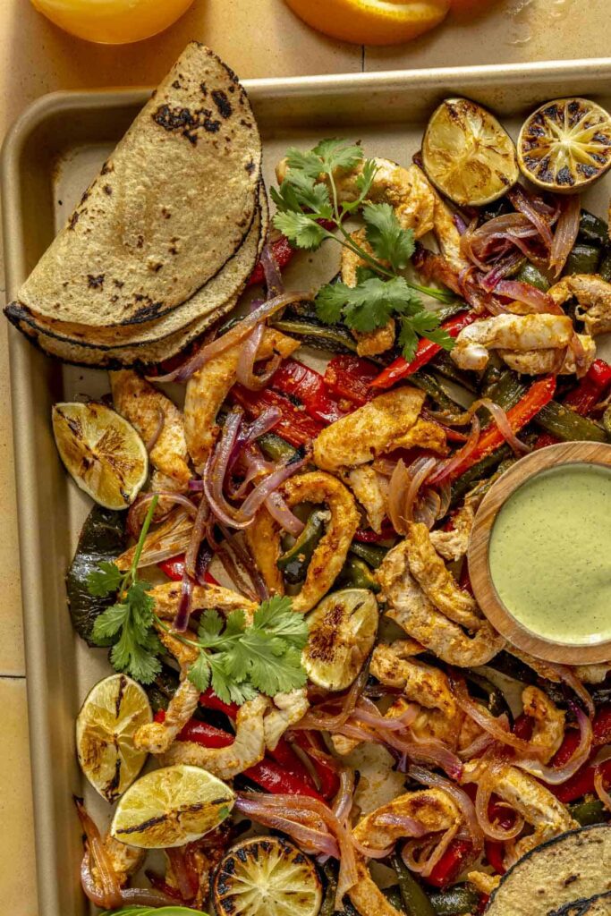 Overhead view of baked fajitas on a baking sheet with vibrant veggies and sauce for dipping.