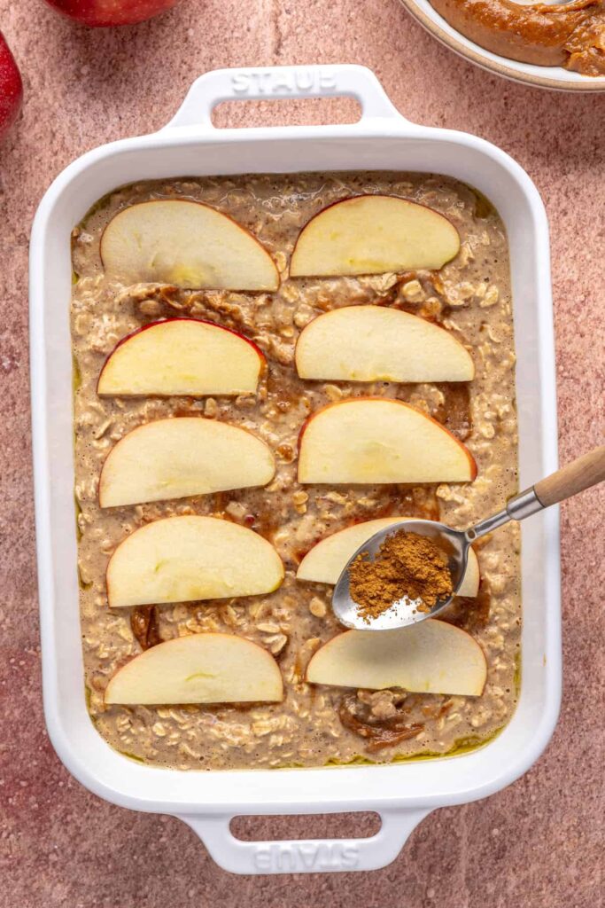 sprinkling cinnamon on top of baked apple oatmeal with caramel swirled throughout and sliced apples on top, ready to bake in a baking dish