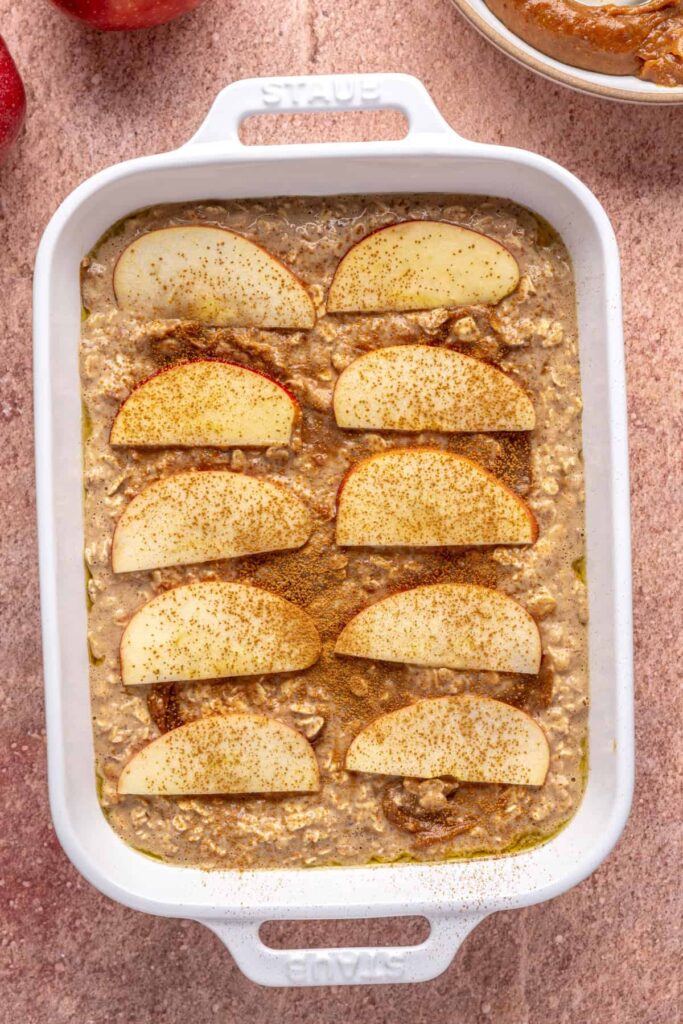 Baked apple oatmeal with caramel swirled throughout and sliced apples and cinnamon sprinkled on top, ready to bake in a baking dish