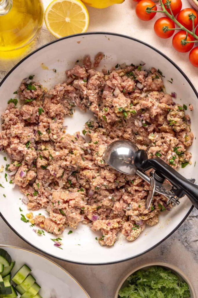 Scooping ground turkey meatball mixture with herbs, garlic, and breadcrumbs in a bowl for Greek turkey meatballs.