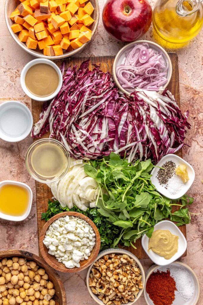 Overhead view of ingredients for ultimate harvest salad: radicchio, frisée, fennel, apples, sweet potatoes, chickpeas, walnuts, feta, and tahini dressing
