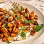 Drizzling hot honey over roasted sweet potatoes on a plate with whipped feta and toasted nuts