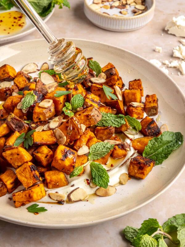 Drizzling hot honey over roasted sweet potatoes on a plate with whipped feta and toasted nuts