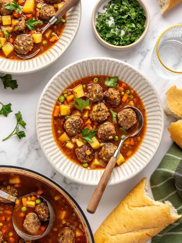 Overhead view of beef meatball soup served in a bowl with crusty baguette and on a table with garnishes