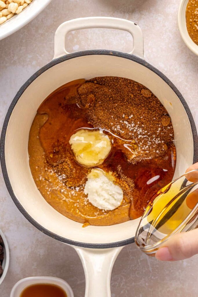 Pouring coconut oil into a pot with peanut butter, maple syrup, coconut sugar, and salt for salted peanut butter bars.