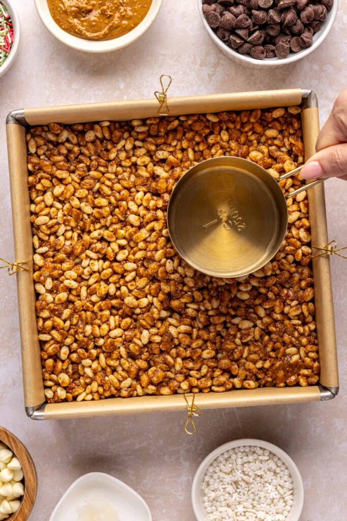 Pressing salted peanut butter bars base into an 8x8 parchment-lined pan with a measuring cup.