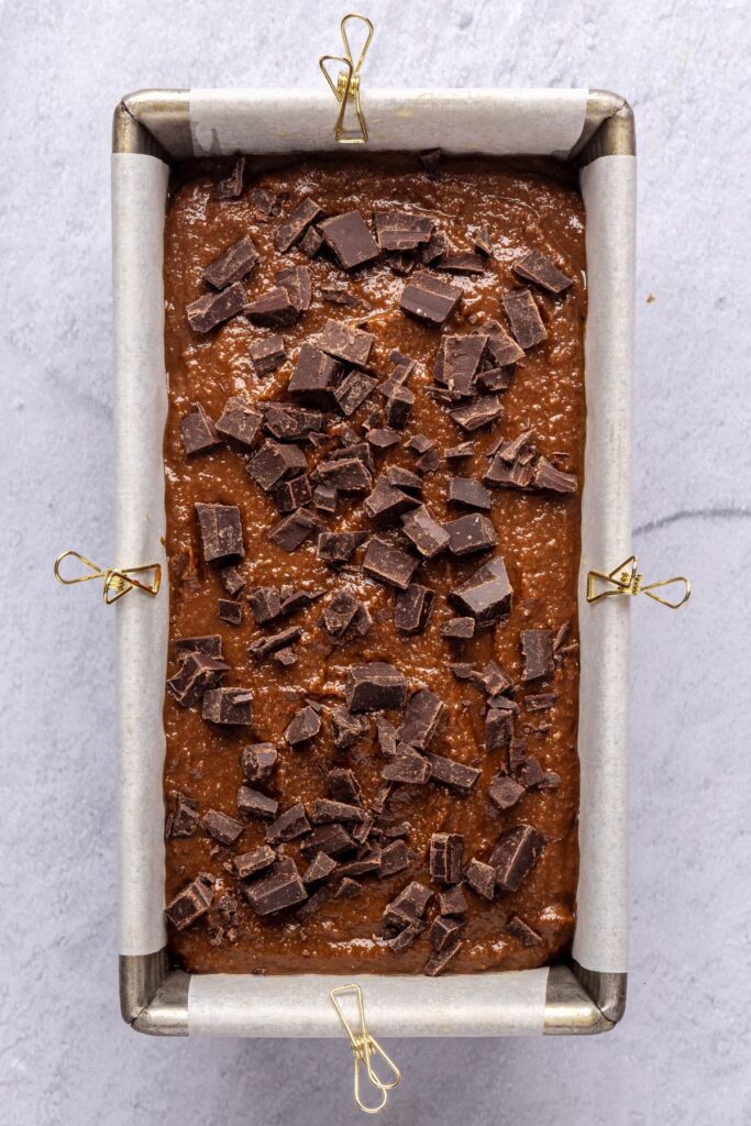 Pumpkin spice chocolate loaf batter poured into a lined loaf pan and topped with chopped dark chocolate before baking.