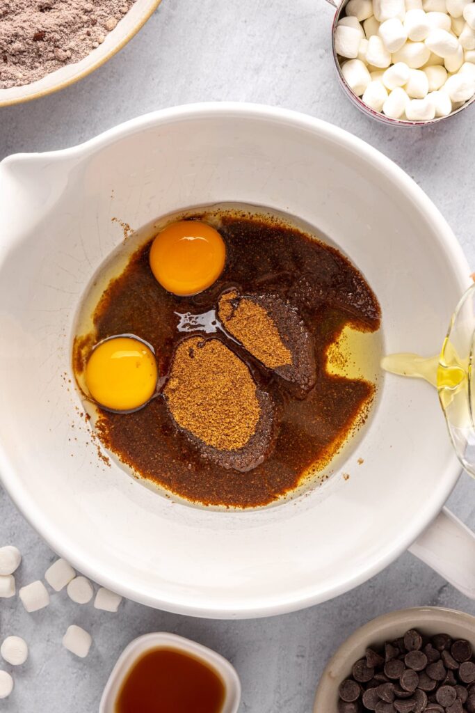 pouring avocado oil into the wet ingredients for hot cocoa cookies, including eggs, coconut sugar, in a large mixing bowl