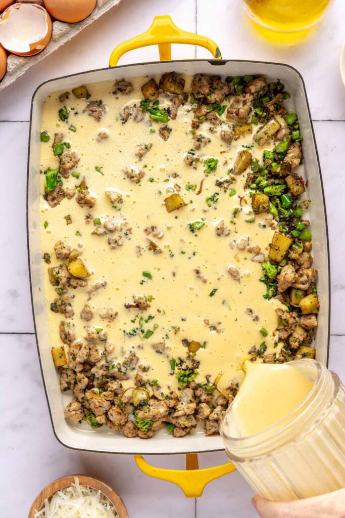 Blended egg mixture being poured over sausage and vegetables in baking dish