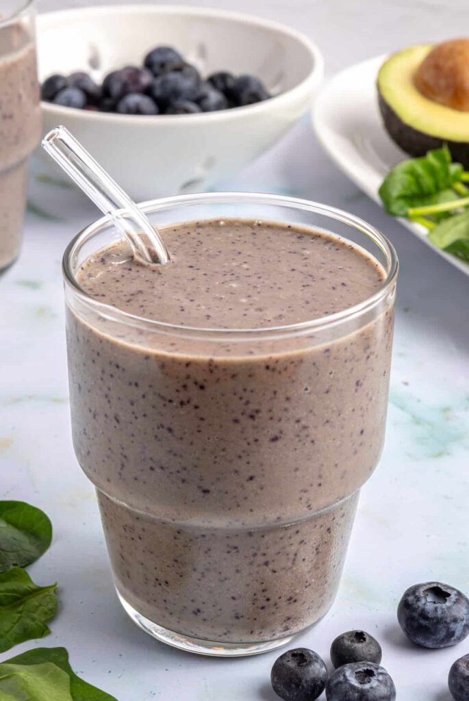 Toddler smoothie served in a glass with a reusable glass straw, on a table styled with blueberries and other ingredients nearby