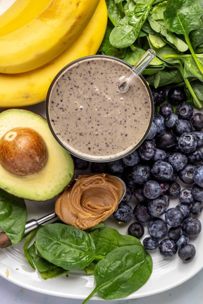 Overhead close-up of toddler smoothie in glass with banana, blueberries, spinach, peanut butter, and avocado arranged around it
