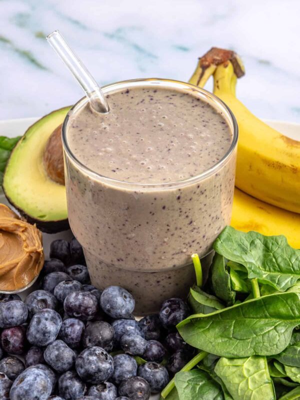 Toddler smoothie in a glass on a table surrounded by fresh banana, blueberries, spinach, peanut butter, and avocado