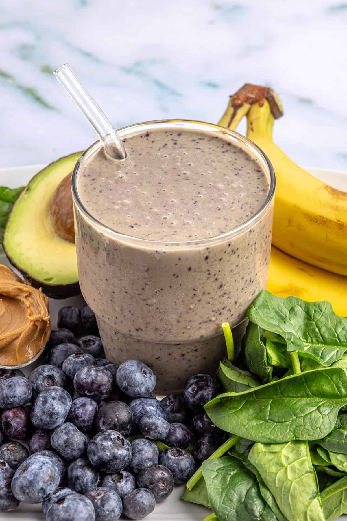 Toddler smoothie in a glass on a table surrounded by fresh banana, blueberries, spinach, peanut butter, and avocado