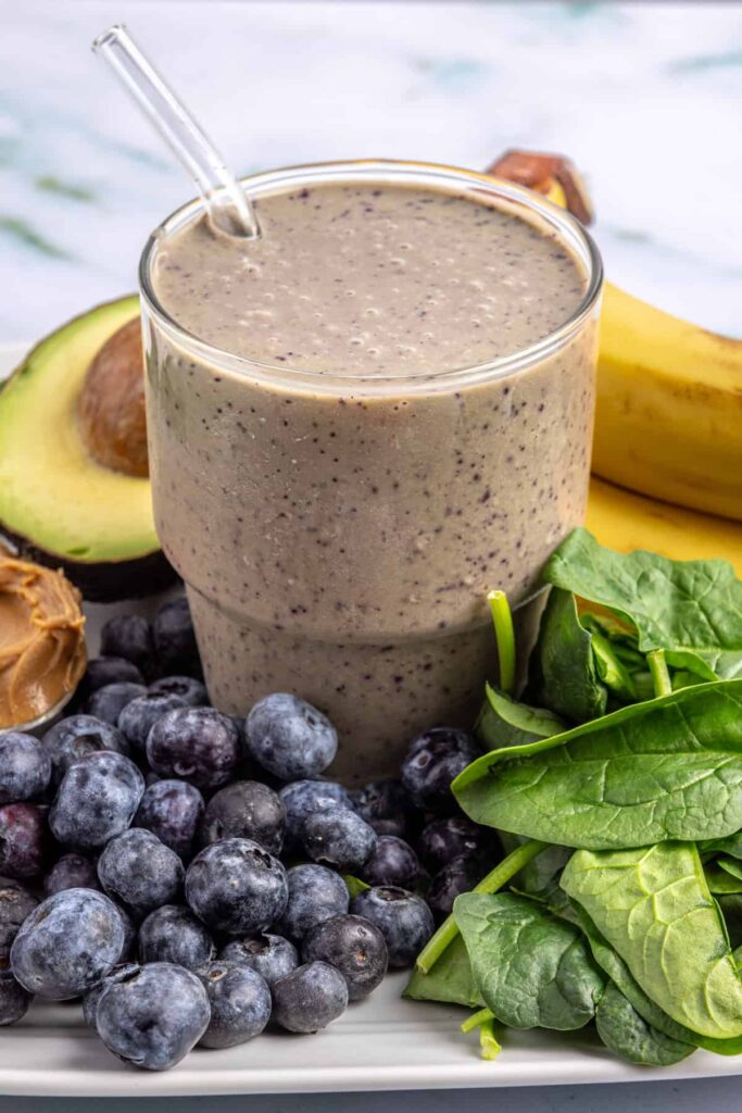 Toddler smoothie in a glass on a table surrounded by fresh banana, blueberries, spinach, peanut butter, and avocado