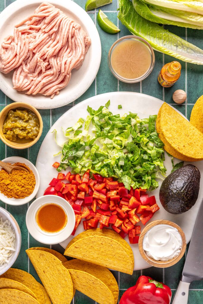 Overhead view of ingredients for easy turkey tacos, including ground turkey, taco seasoning, green chiles, tortillas, romaine, bell pepper, and avocado