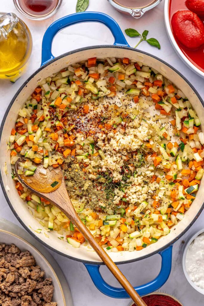 Vegetables cooking down in a Dutch oven, with seasonings being stirred in, for creamy vodka sauce with hidden veggies