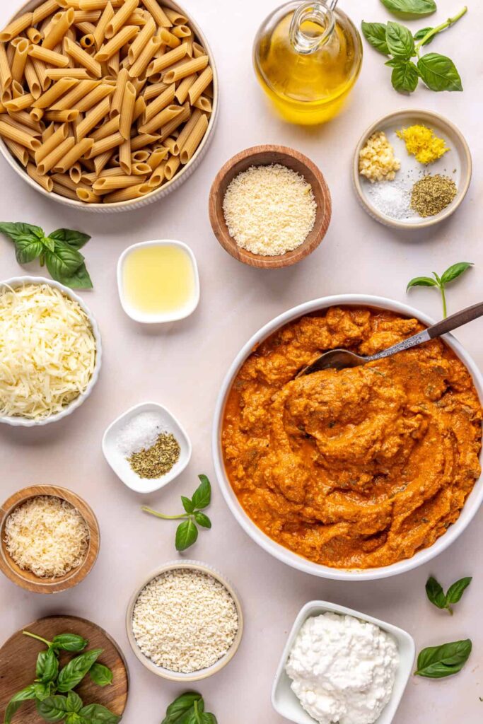Overhead view of ingredients for easy baked ziti, including brown rice pasta, vodka sauce, cottage cheese, mozzarella, parmesan, basil, and gluten-free panko