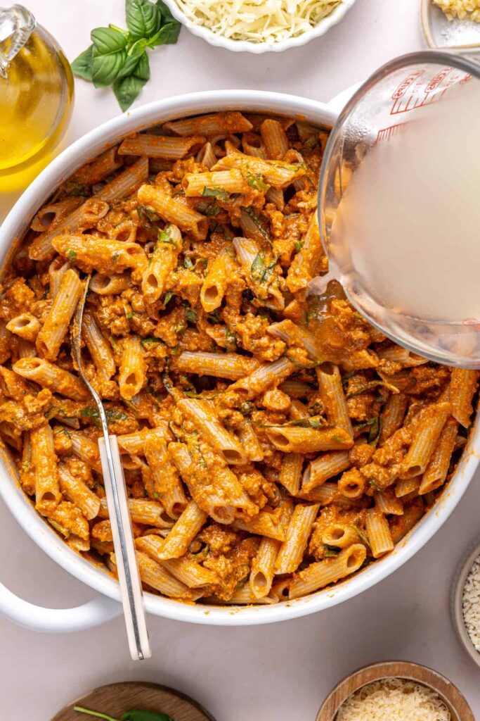 adding pasta water to the bowl of pasta coated in homemade vodka sauce and fresh basil before assembling easy baked ziti
