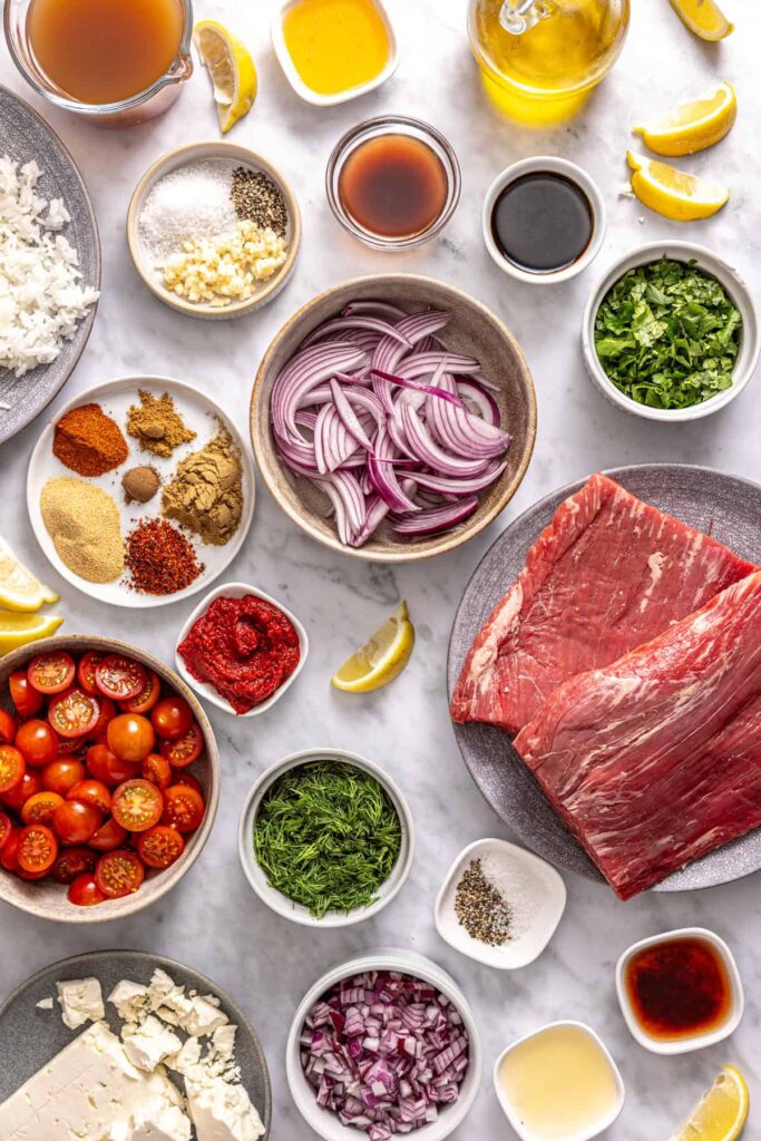 Overhead shot of ingredients for Greek beef bowls including flank steak, spices, tomatoes, herbs, and lemon
