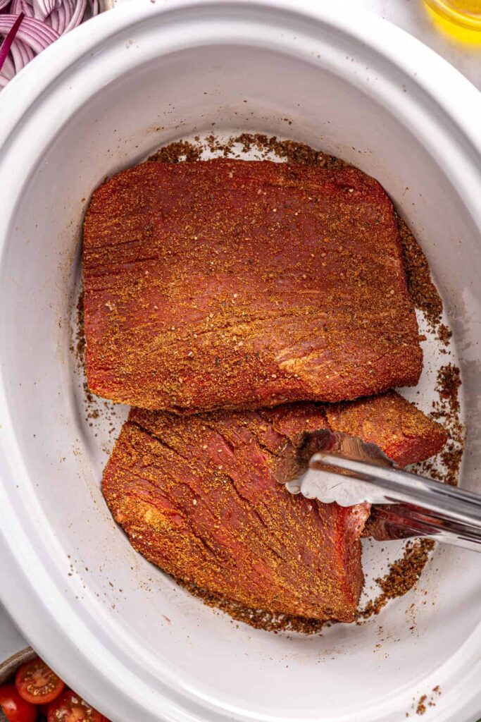 Flank steak seasoned with Greek spice blend being placed into a slow cooker with tongs
