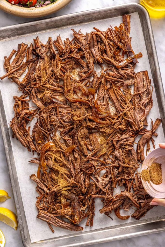 adding the remaining spice blend to the shredded flank steak on a sheet pan