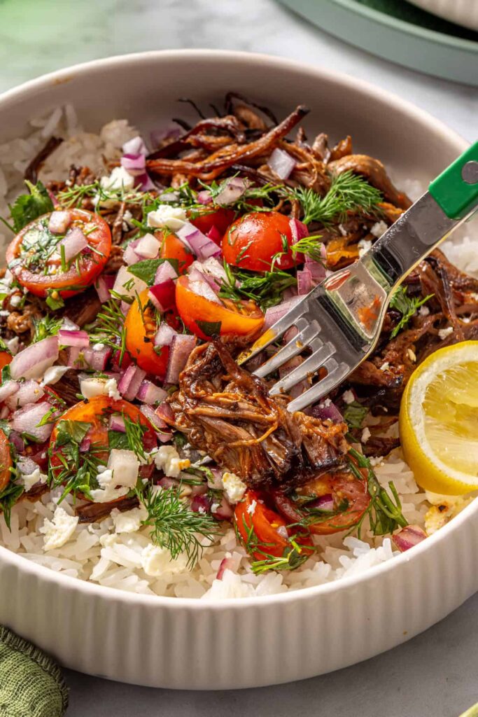 close up photo of Slow Cooker Crispy Greek Beef Bowls with rice, tomato relish, feta, and lemon wedge, served with a fork