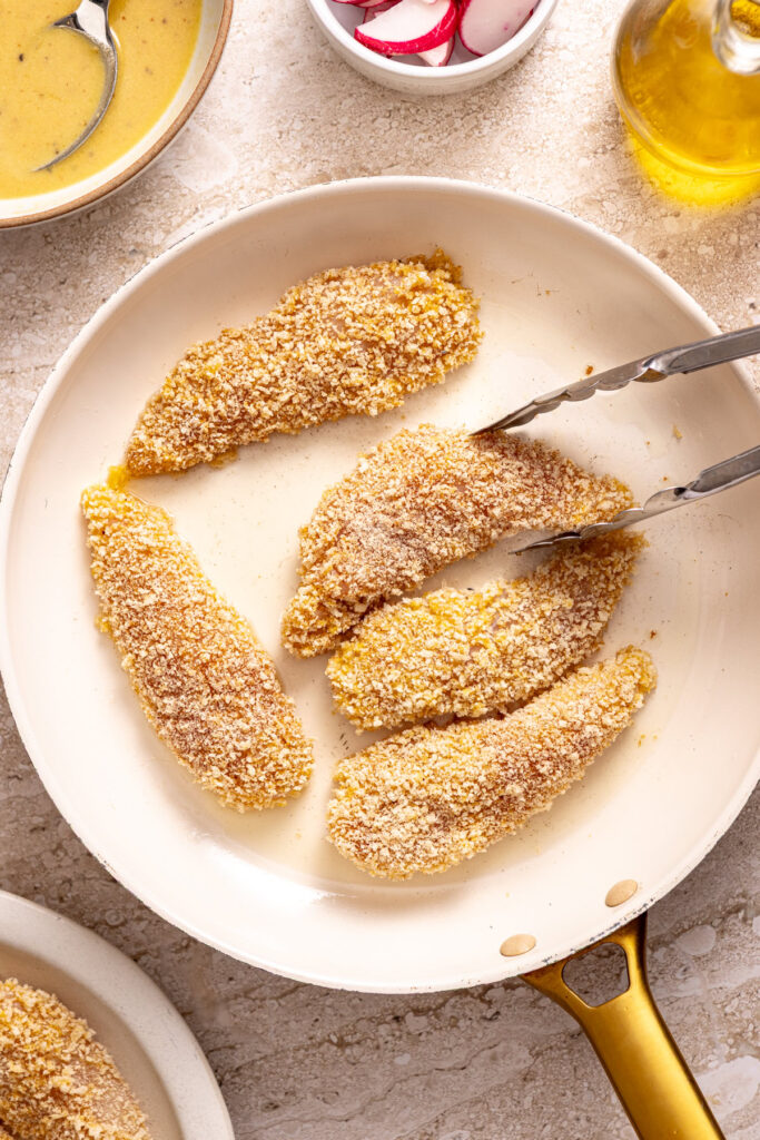 chicken tenders coated in gluten-free panko breadcrumbs being transferred with tongs to a baking sheet
