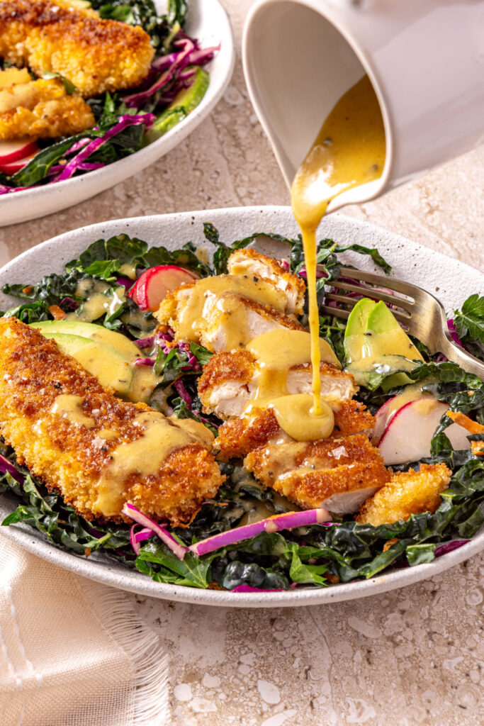 White ceramic pitcher pouring honey mustard dressing onto chicken kale salad