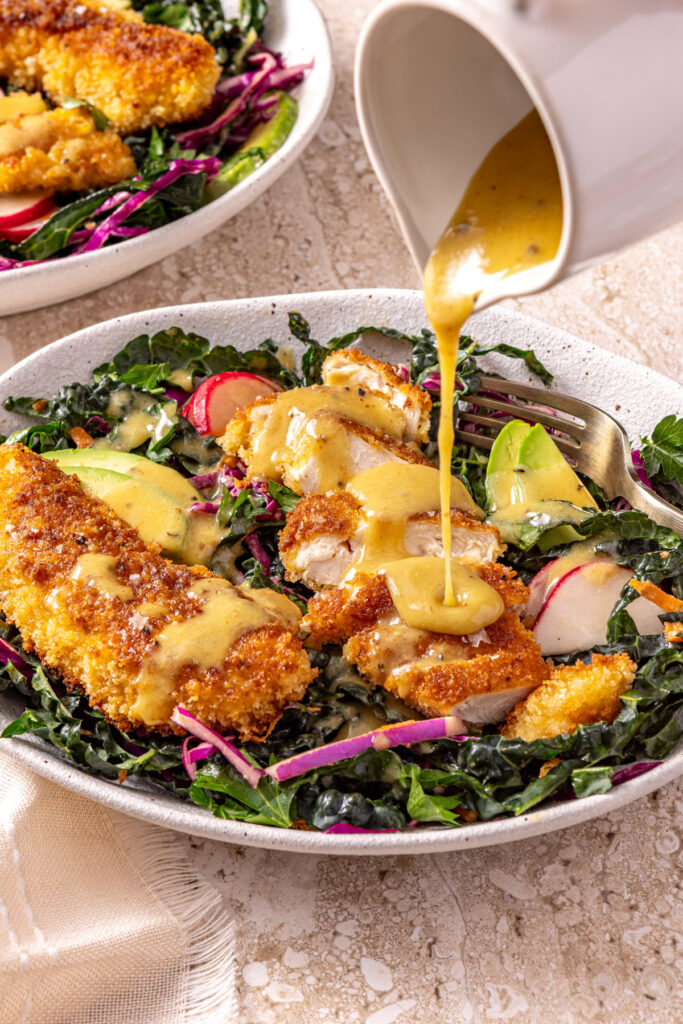Honey mustard dressing being poured over crispy chicken kale salad