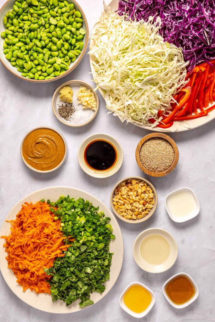 Overhead shot of ingredients for crunchy peanut cabbage salad including green cabbage, red cabbage, carrots, edamame, peanut butter, and sesame oil