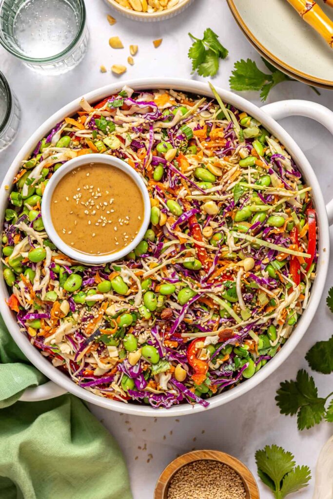 Crunchy peanut cabbage salad served in a large white bowl with small bowl of dressing and peanuts on styled table