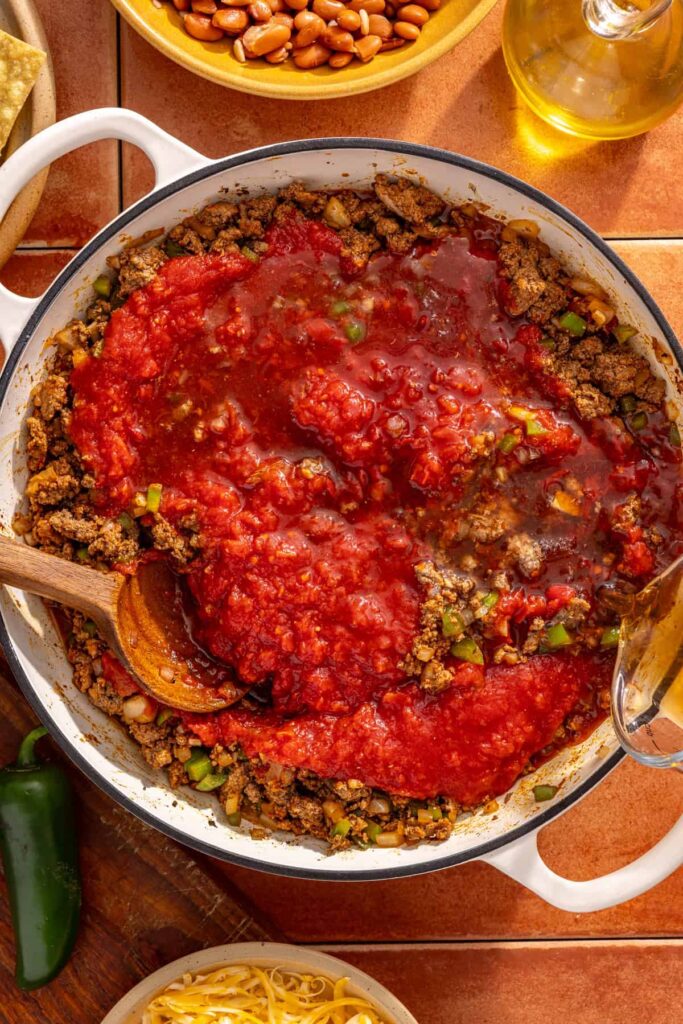 Adding crushed tomatoes and broth to chili skillet