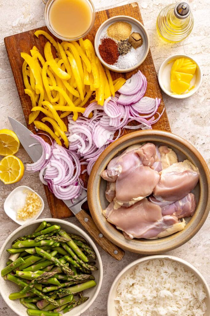 Ingredients for chicken thigh rice bowls including chicken thighs, asparagus, peppers, onion and spices overhead