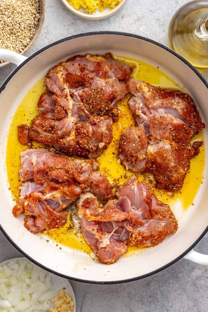 Chicken thighs searing in a large skillet for lemon herb chicken quinoa pilaf