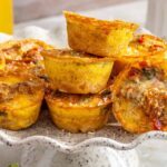 Styled overhead shot of gluten-free quiche cups stacked on a cake stand with some turned on their side to show the layered potato crust and fluffy egg filling, served with fresh fruit and breakfast sides