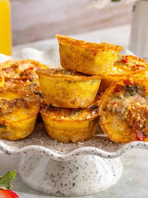 Styled overhead shot of gluten-free quiche cups stacked on a cake stand with some turned on their side to show the layered potato crust and fluffy egg filling, served with fresh fruit and breakfast sides