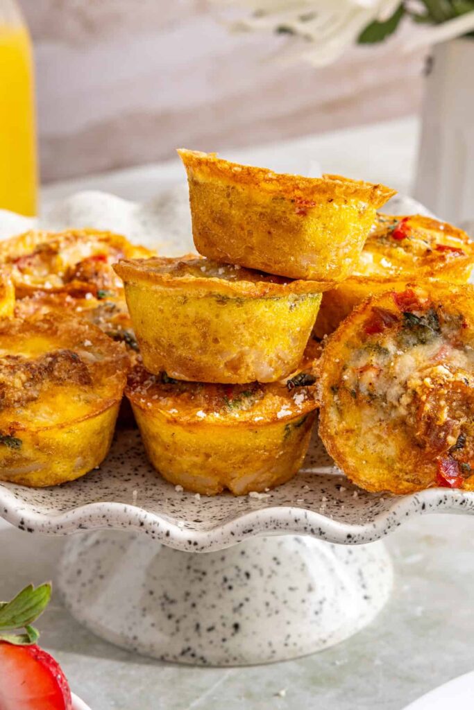 Styled overhead shot of gluten-free quiche cups stacked on a cake stand with some turned on their side to show the layered potato crust and fluffy egg filling, served with fresh fruit and breakfast sides