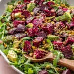Roasted beet salad with goat cheese, avocado, arugula, pistachios, and tahini dressing served in a platter with a serving spoon on a styled table