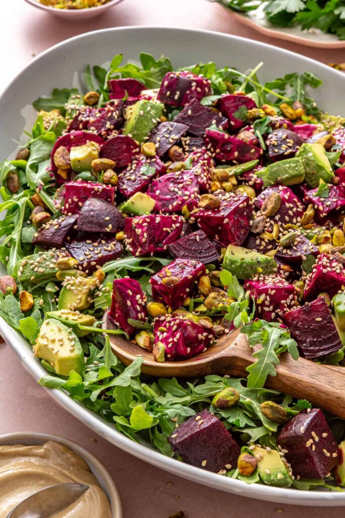 Roasted beet salad with goat cheese, avocado, arugula, pistachios, and tahini dressing served in a platter with a serving spoon on a styled table