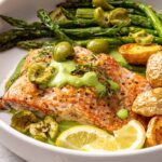 Baked sheet pan salmon and asparagus served in a bowl with herb yogurt sauce, olives, potatoes, and lemon wedges