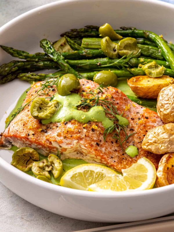 Baked sheet pan salmon and asparagus served in a bowl with herb yogurt sauce, olives, potatoes, and lemon wedges