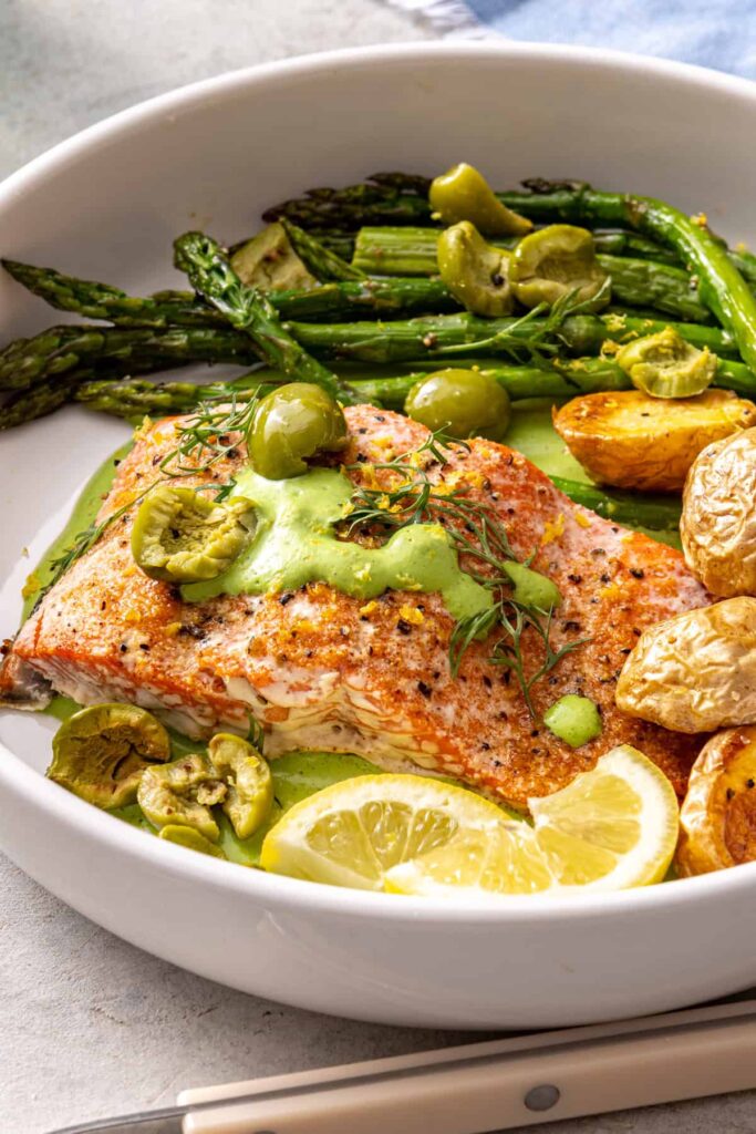 Baked sheet pan salmon and asparagus served in a bowl with herb yogurt sauce, olives, potatoes, and lemon wedges