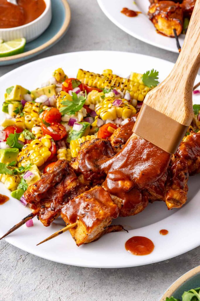 Brushing BBQ sauce onto grilled chicken skewers served with corn and tomato salad