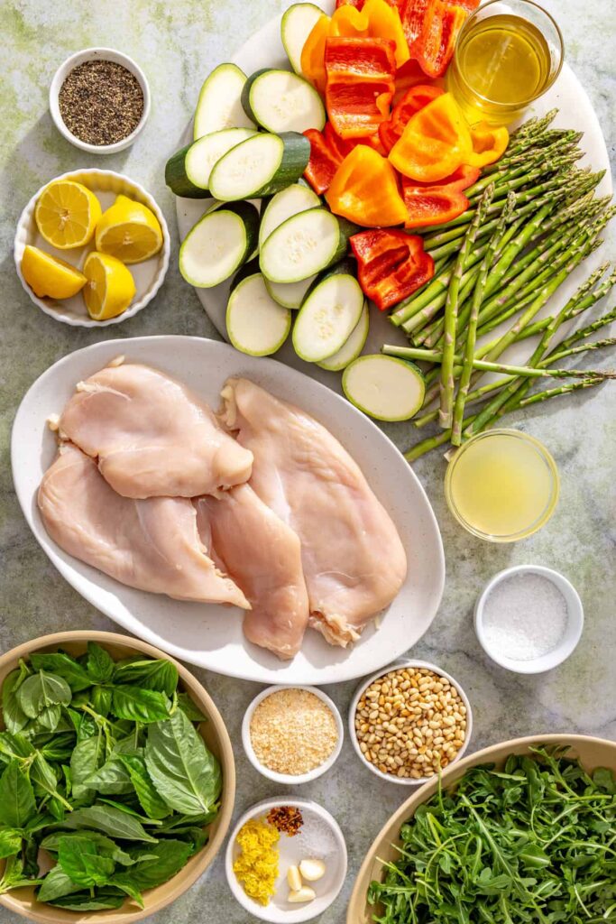 Ingredients for lemon pesto grilled chicken including chicken breasts, basil, pine nuts, lemons, zucchini, asparagus, bell peppers, garlic, Parmesan, and olive oil