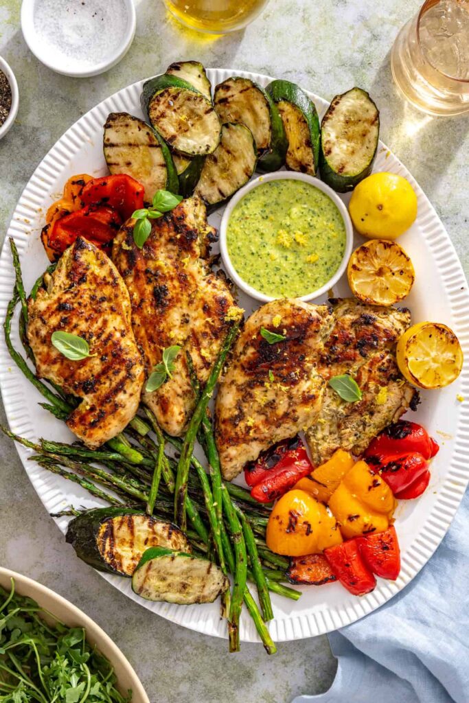 Overhead table setting featuring lemon pesto grilled chicken, grilled vegetables, salad, and summer drinks