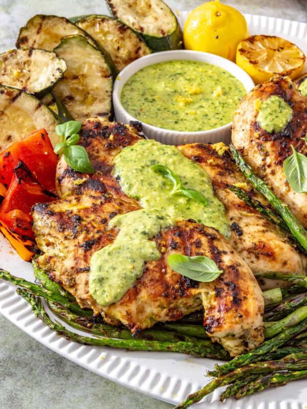 Lemon pesto grilled chicken served with grilled zucchini, asparagus, and bell peppers on a white serving platter, topped with homemade pesto and fresh lemon