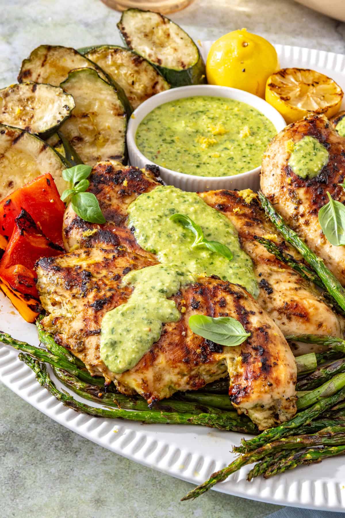 Lemon pesto grilled chicken served with grilled zucchini, asparagus, and bell peppers on a white serving platter, topped with homemade pesto and fresh lemon