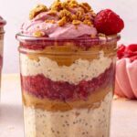 Peanut butter and jelly overnight oats in a glass jar topped with raspberry Greek yogurt whip, peanut butter oat crumble, and a fresh raspberry