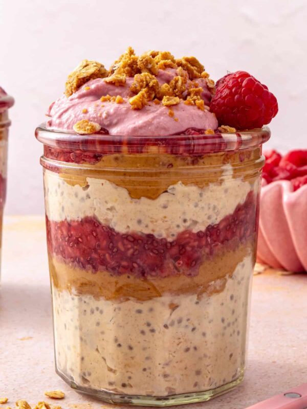 Peanut butter and jelly overnight oats in a glass jar topped with raspberry Greek yogurt whip, peanut butter oat crumble, and a fresh raspberry