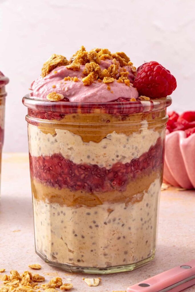 Peanut butter and jelly overnight oats in a glass jar topped with raspberry Greek yogurt whip, peanut butter oat crumble, and a fresh raspberry